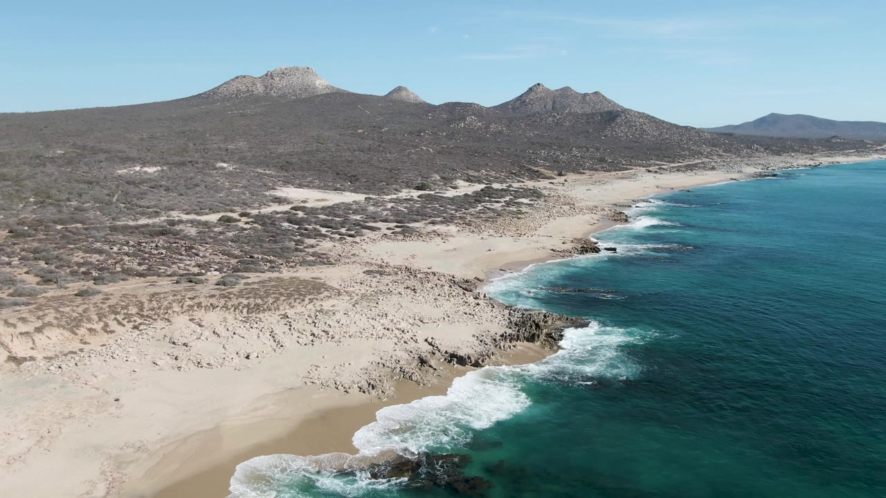 Buzzards Beach coastline from above with a band of white waves curling onto sand, dark rocks near the shore, dry scrubland inland, and no visible people.
