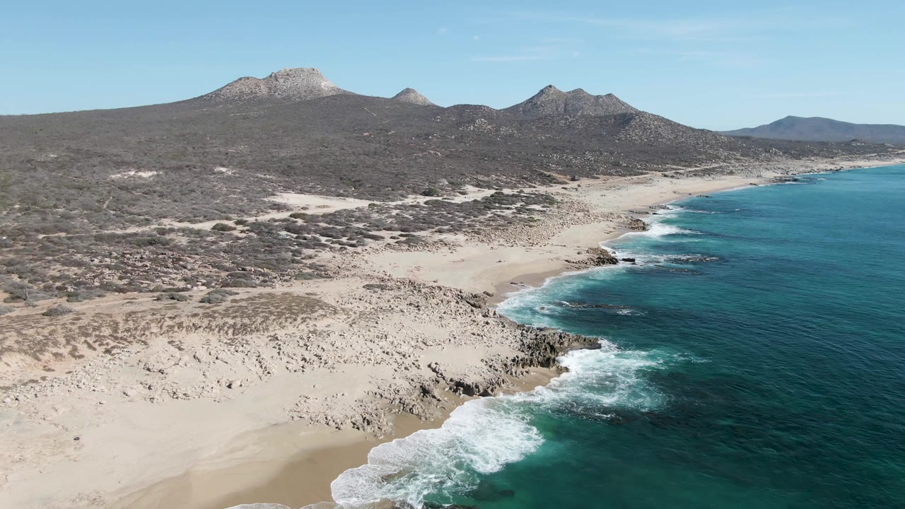 Aerial frame of the curving Buzzards Beach shoreline, with clear blue-green water on the right, pale sand below, low mountains inland, and no visible people.