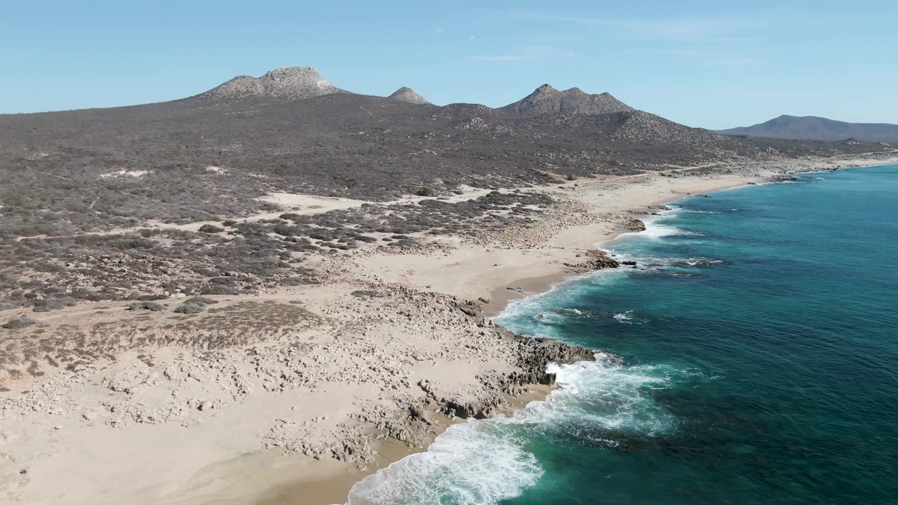 High view over Buzzards Beach showing foamy waves in the lower right, wide pale sand to the left, rocky points along the waterline, inland scrub, and no visible people.
