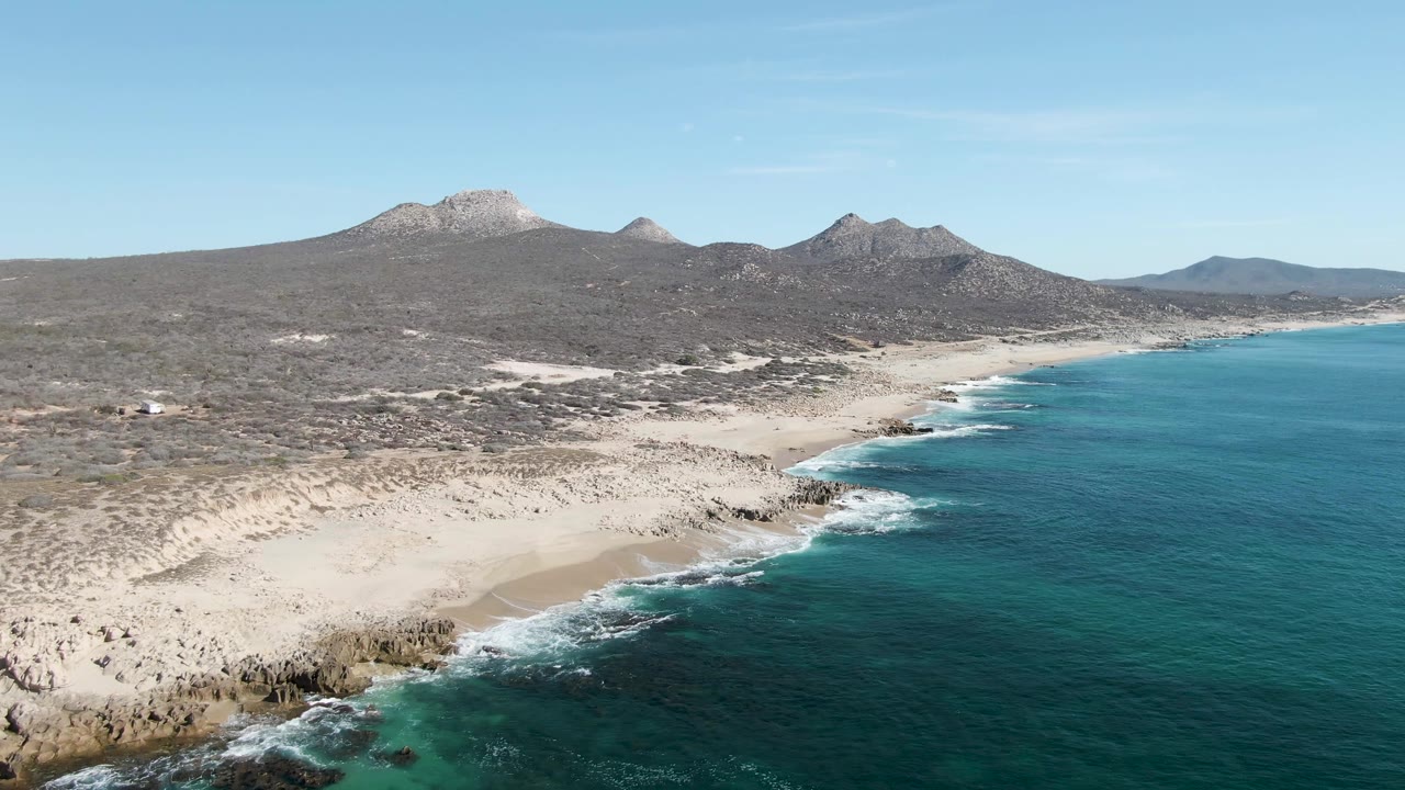 Aerial frame over Buzzards Beach, Baja California Sur, with turquoise water on the right, a sandy cove and rocky shoreline below, inland desert scrub, distant mountains, and no visible people.