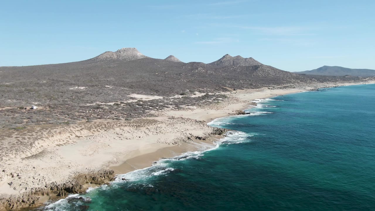 A second aerial view of Buzzards Beach shows the shoreline angled through frame, with white waves breaking on rocks, pale sand inland, blue ocean to the right, mountain ridges beyond, and no visible people.