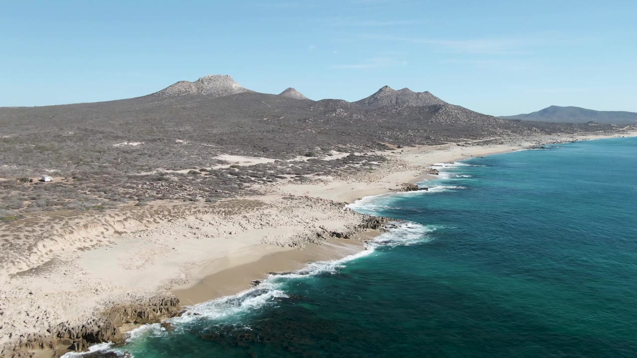 The aerial composition over Buzzards Beach holds the empty sand near the lower left, darker rocks at the waterline, turquoise ocean across the right half, desert scrub behind the beach, and no visible people.