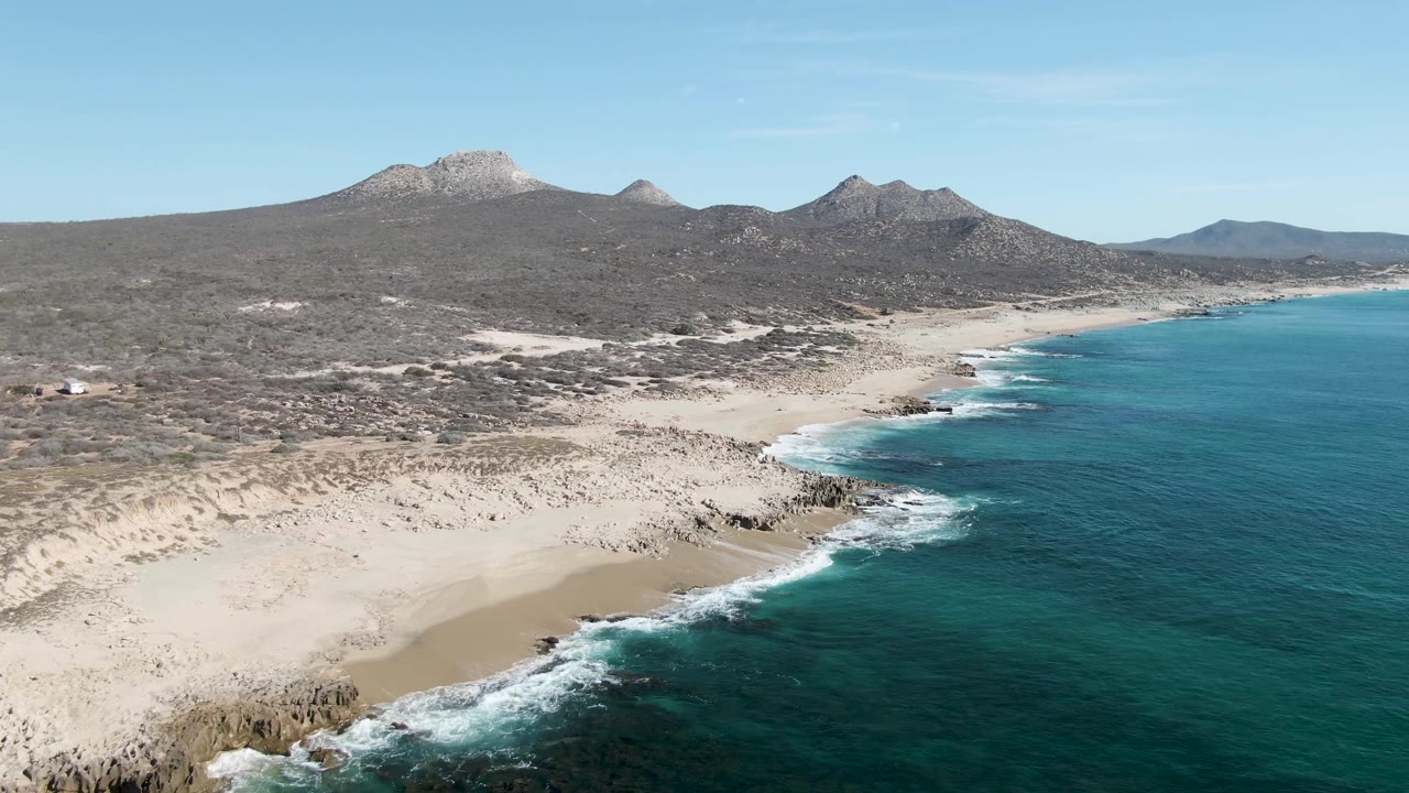 Aerial coastline with sandy foreground, surf, ocean, and mountains