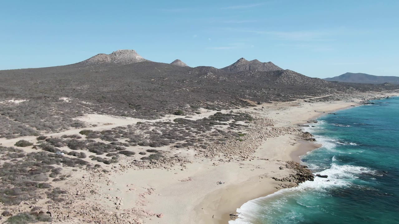 Aerial view of sandy beach, turquoise surf, desert scrub, and mountains.