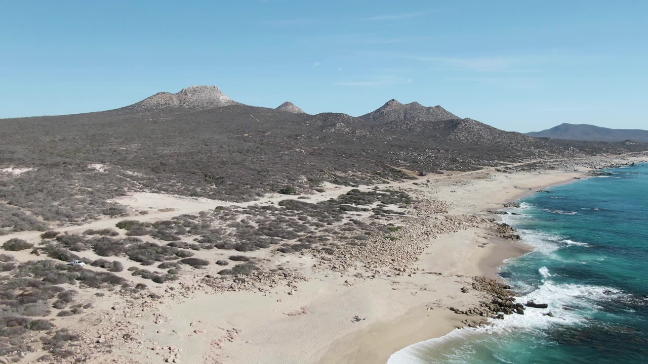 Wide aerial view at Buzzards Beach with the surf line shifted slightly along the right edge, pale sand in the foreground, low shrubs inland, mountains behind, and no visible people.