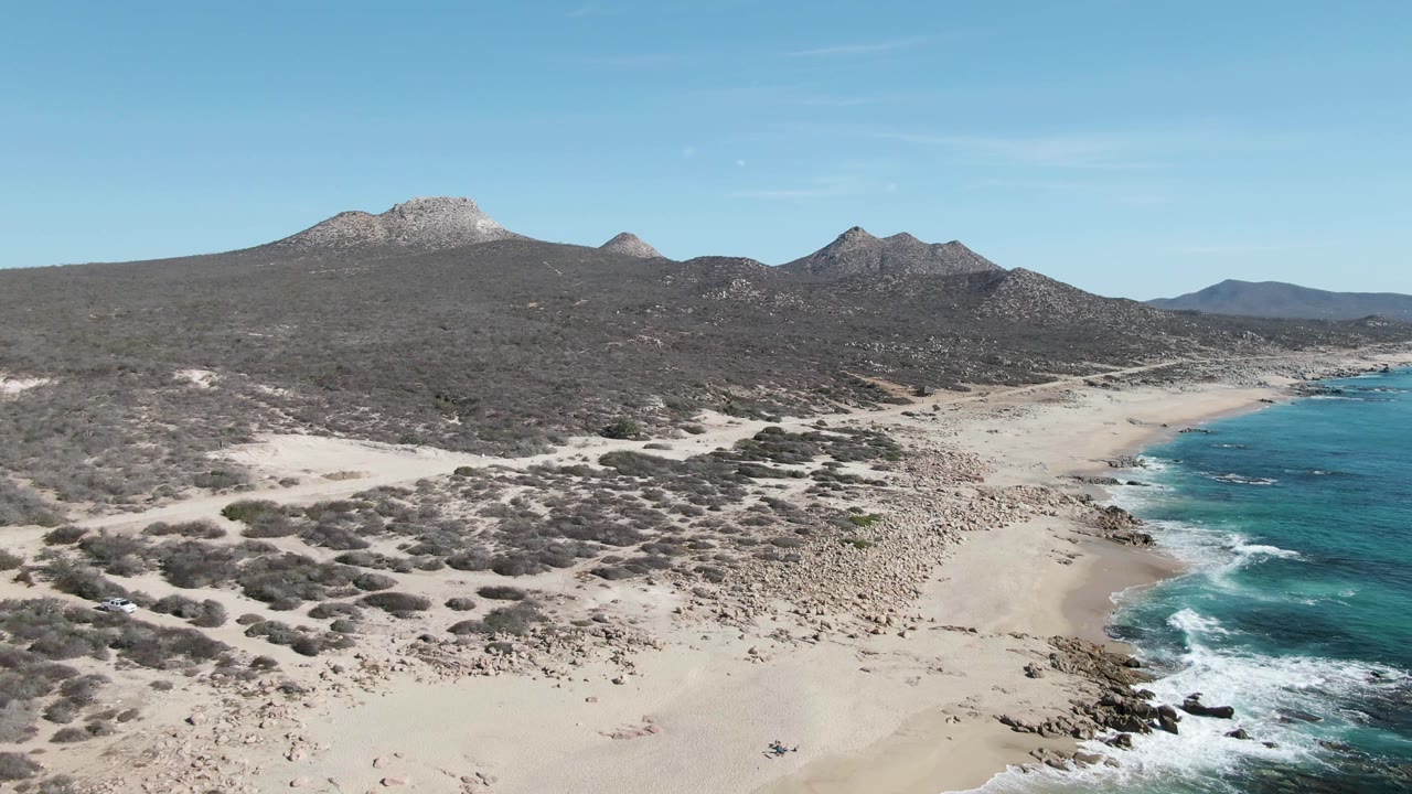 Aerial frame from Buzzards Beach showing a broader curve of turquoise water and white surf on the right, open sand and rocky beach in the center, scrubland beyond, and no visible people.