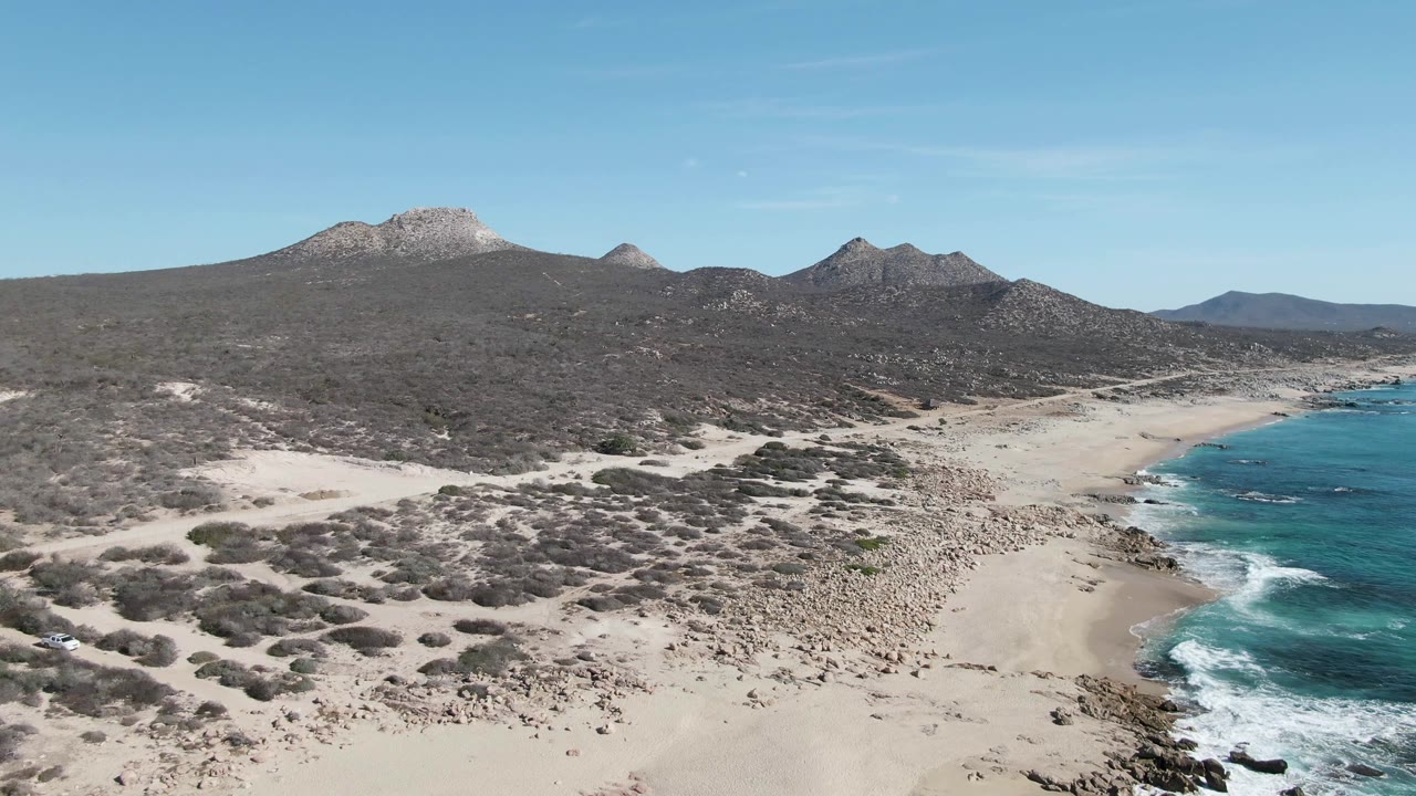 Later aerial frame from Buzzards Beach with more foreground scrub visible, pale sand leading toward the surf at right, rugged mountains across the background, and no visible people.
