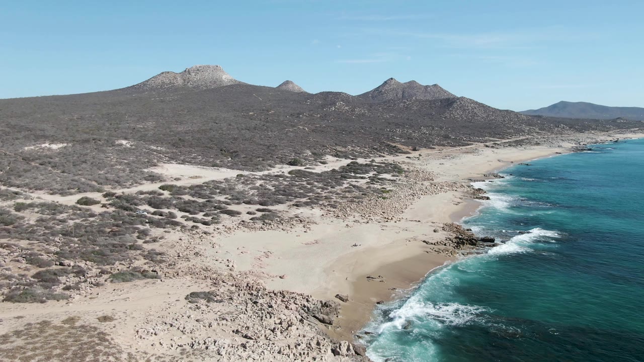 Buzzards Beach appears from an elevated angle with pale sand across the foreground, scattered dark rocks, white waves rolling in from the right, desert scrub, mountains, and no visible people.