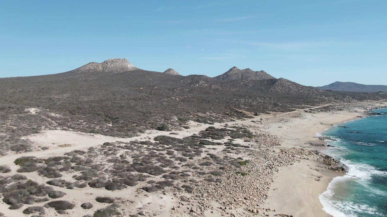 Aerial view of sandy coast, turquoise surf, scrubland, and mountain ridges.