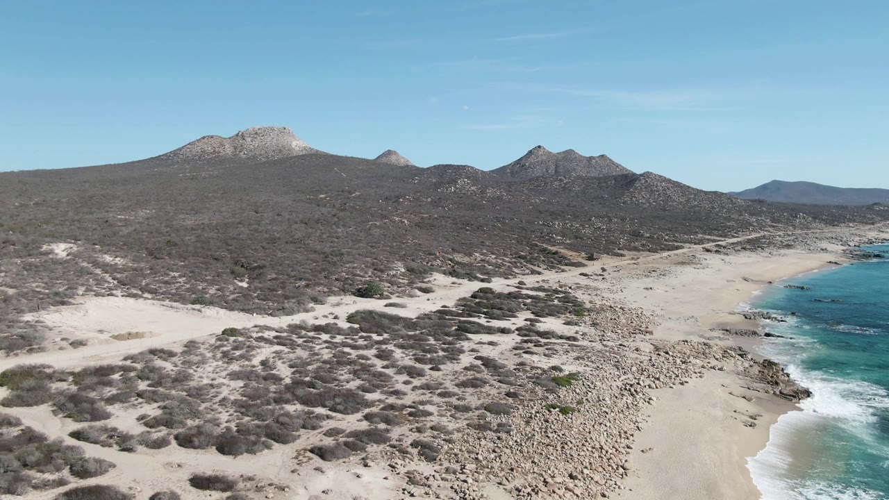 Second frame at Buzzards Beach, Baja California Sur, with scrub-covered sand in the foreground, a curved beach below, turquoise surf along the right edge, mountain ridges beyond, and no visible people.