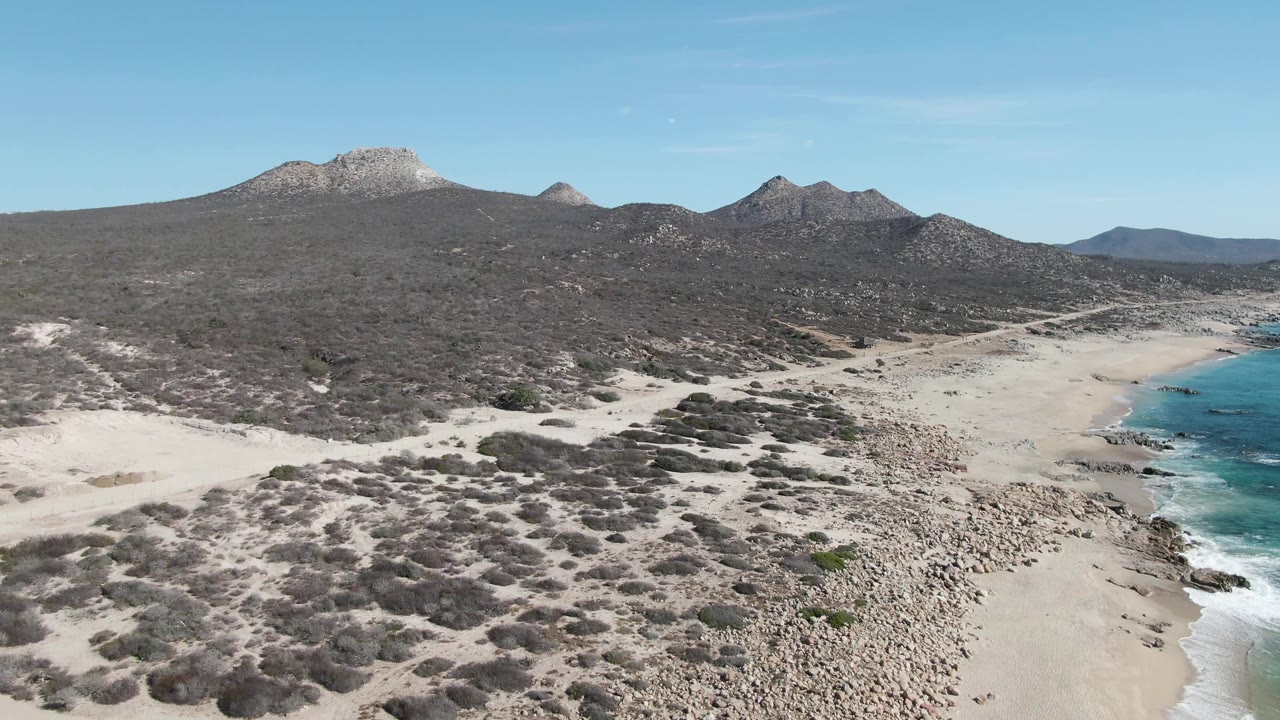 Mid-clip aerial frame at Buzzards Beach, Baja California Sur, with dry scrub filling the foreground, a sandy beach and white surf to the right, dark mountain slopes beyond, and no visible people.