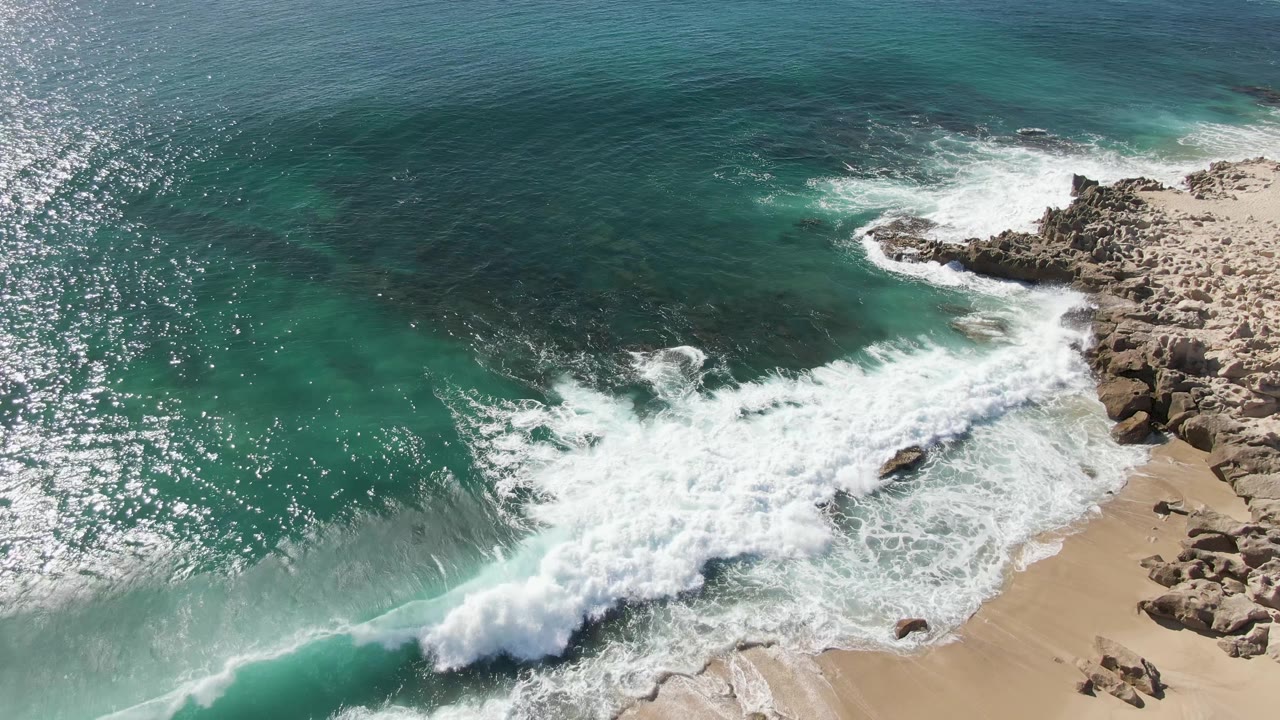 Aerial frame at Buzzards Beach, Baja California Sur, showing turquoise water and white surf rolling onto tan rocks and sand, with no visible people.
