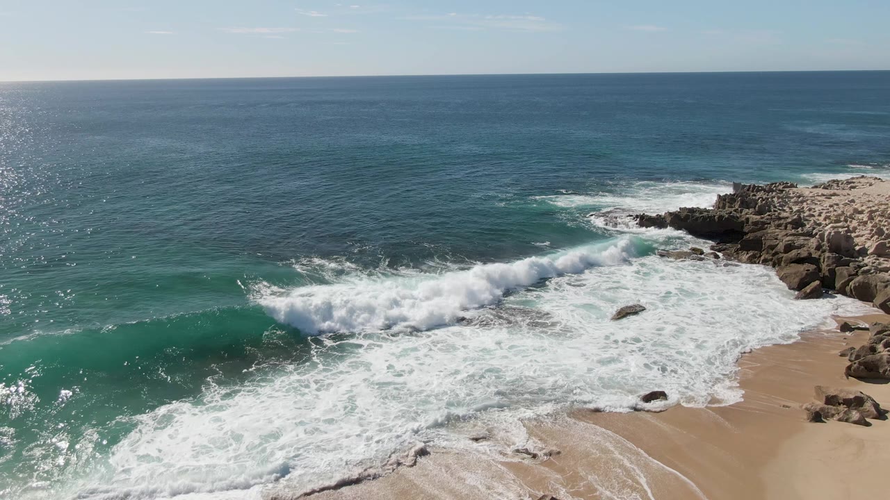 At Buzzards Beach in Baja California Sur, a green-blue wave rolls across the frame toward the rocky right shoreline as foam covers the beach and no people are visible.