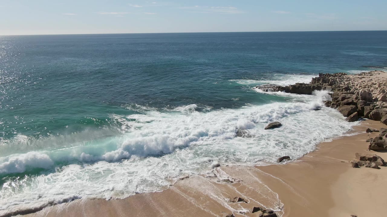 At Buzzards Beach in Baja California Sur, the surf wash pushes across the lower beach with rocks on the right and the open ocean horizon above, with no visible people.