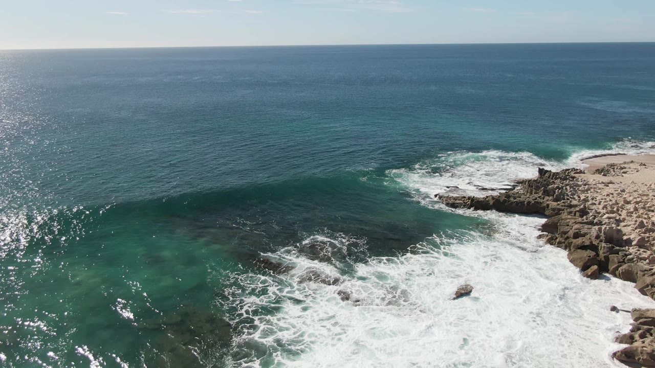 Aerial view of waves breaking over rocks beside turquoise ocean water.