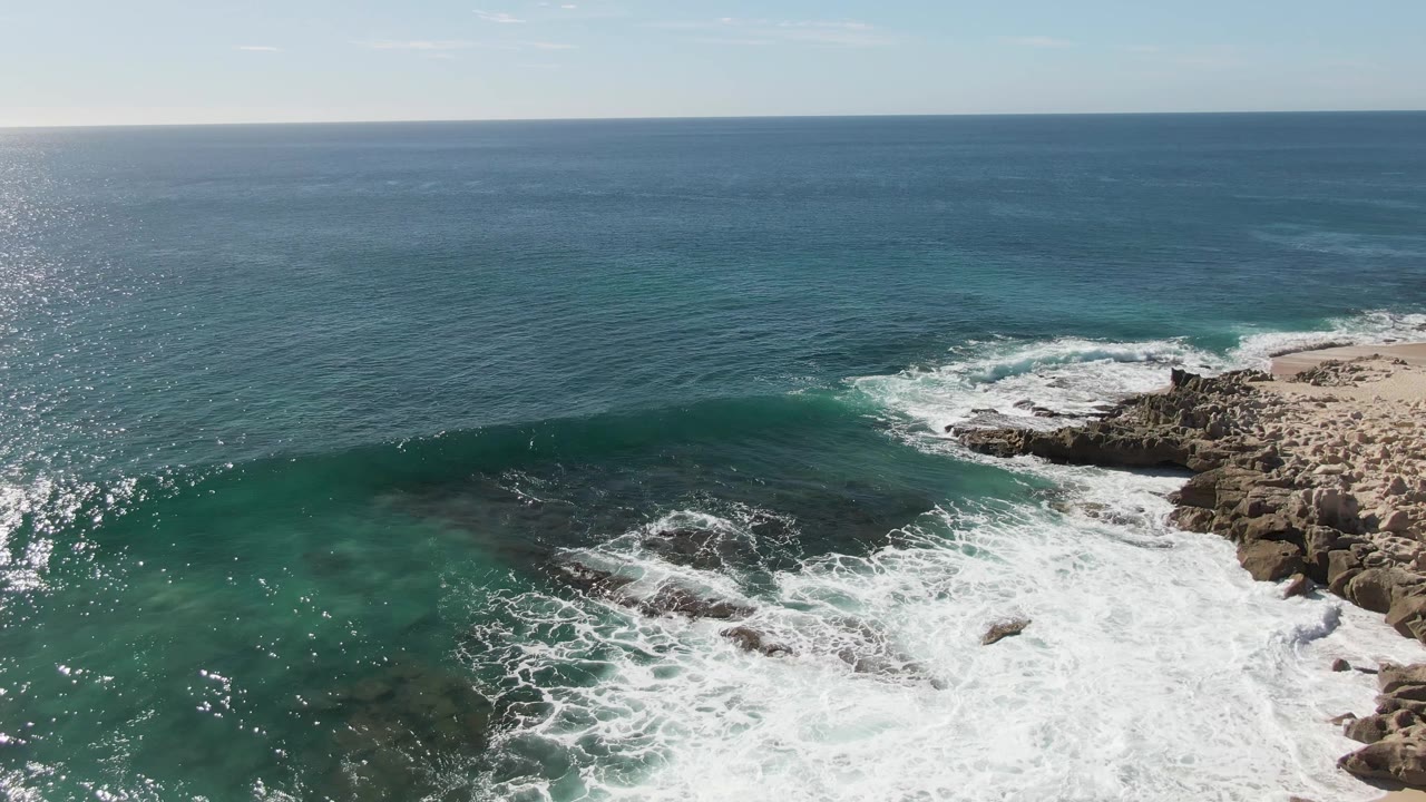 Aerial view at Buzzards Beach with a broad white foam wash over submerged rocks, turquoise shallows in the midground, open blue sea in the background, and no visible people.