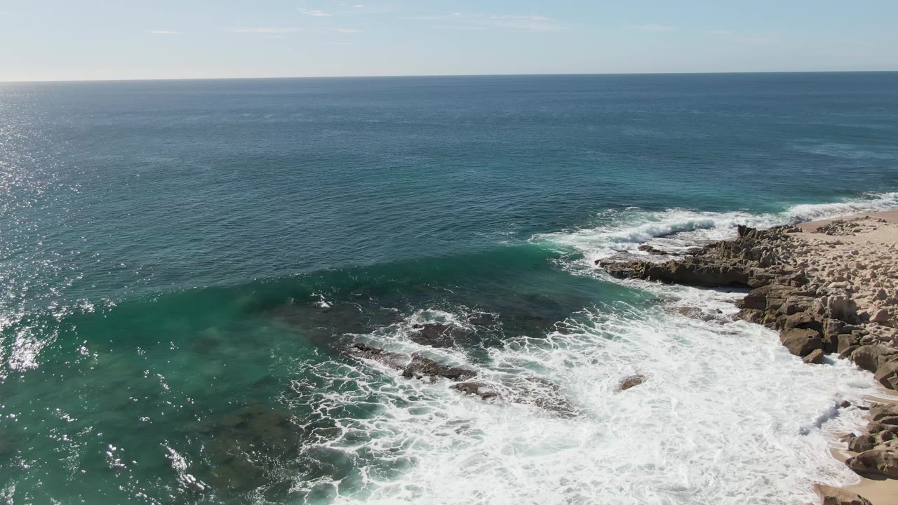 Buzzards Beach aerial frame showing a green-blue swell approaching dark rocks while broken whitewater fills the lower right shoreline and no visible people appear.