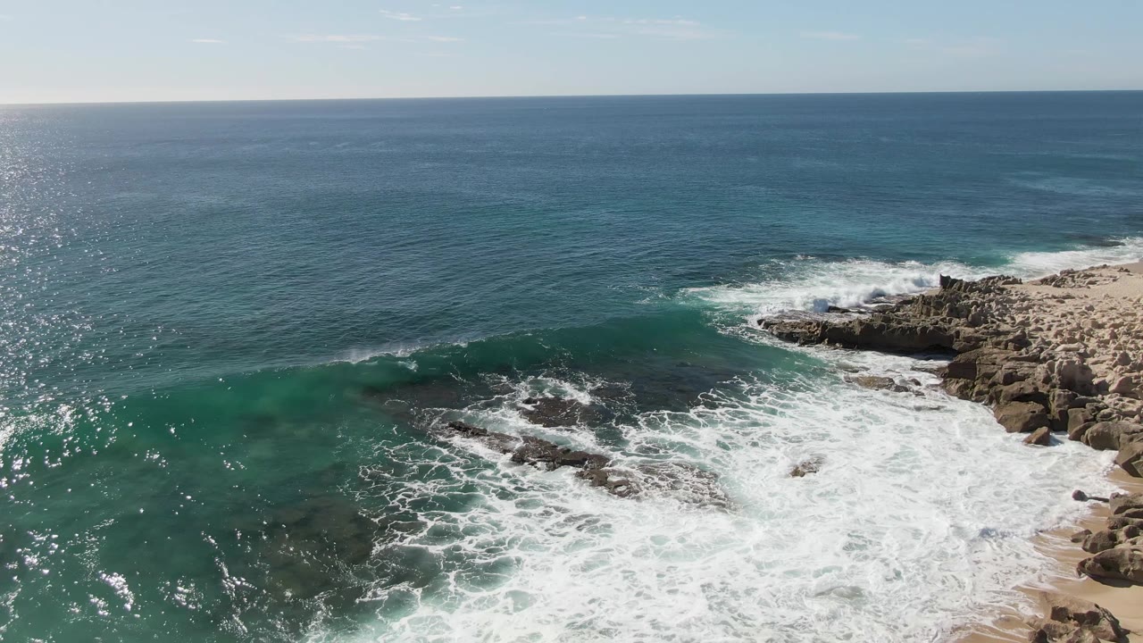 Aerial still from Buzzards Beach with a cleaner turquoise wave band moving across shallow water, white foam below, rocky shoreline to the right, and no visible people.