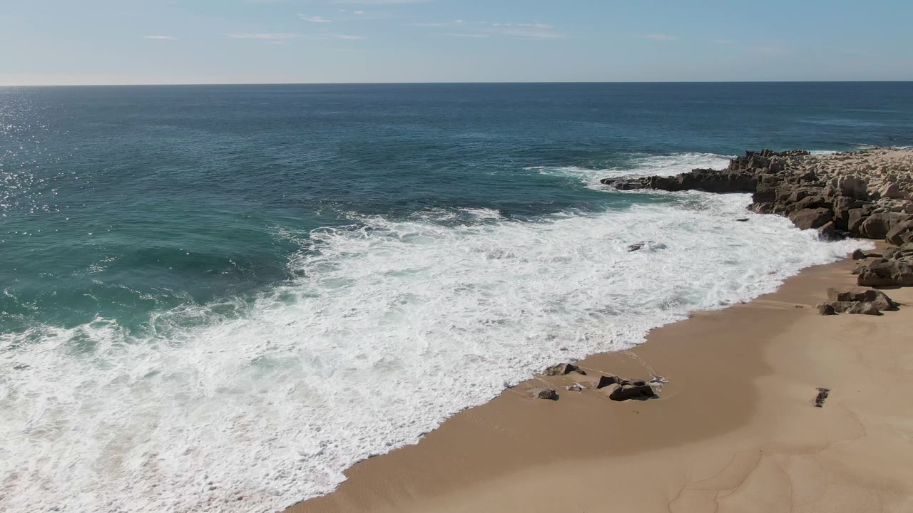 At Buzzards Beach, Baja California Sur, white foam pushes across the sand while turquoise water and right-side rocks remain in view, with no visible people.