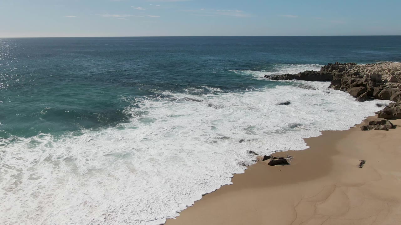Buzzards Beach surf covers much of the lower shoreline, with a blue ocean horizon, tan sand at right, rocky outcrops, and no visible people.