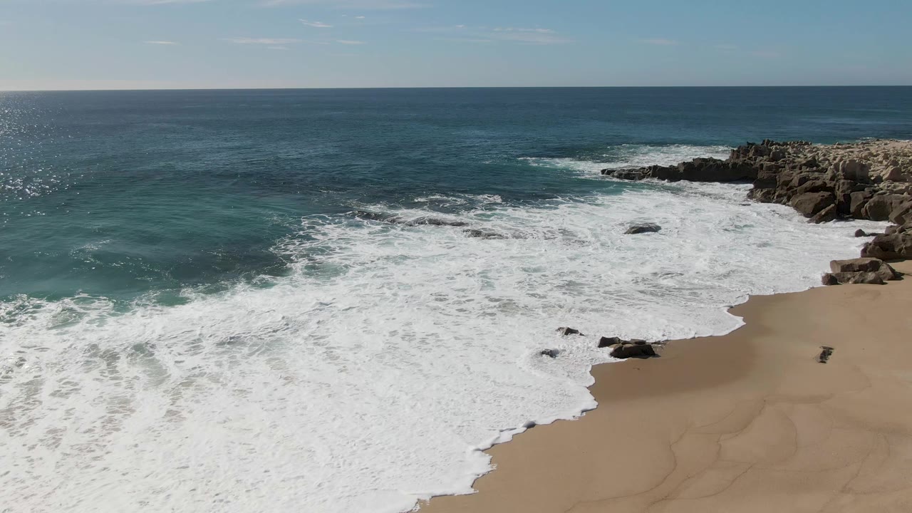 Aerial view at Buzzards Beach with foamy water thinning around beach rocks, open blue ocean beyond, right-side outcrops, and no visible people.