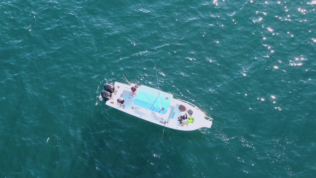 At Buzzards Beach in Baja California Sur, the small boat sits toward the upper right of the aerial frame, with people visible near the bow and stern and bright flecks on the water.