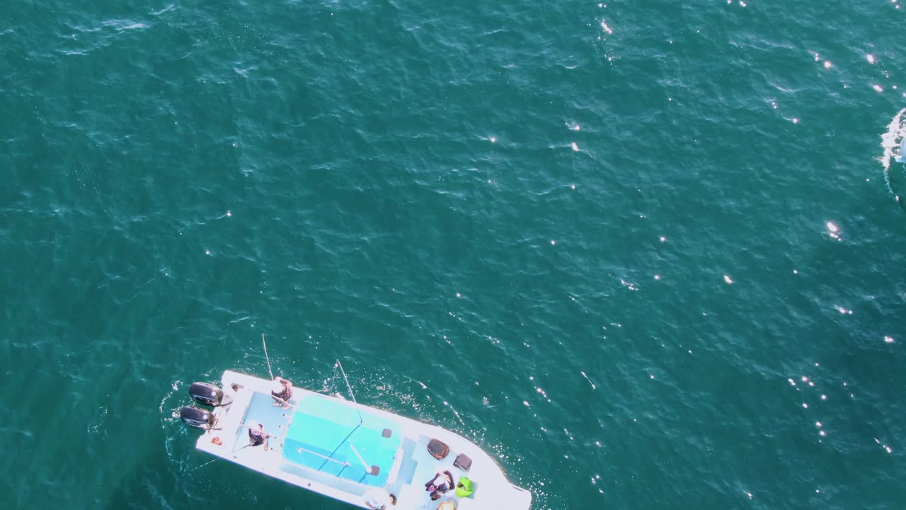 Top-down drone view at Buzzards Beach showing the boat slightly farther right in the frame, with people visible on deck and bright points of sunlight on textured green-blue water.