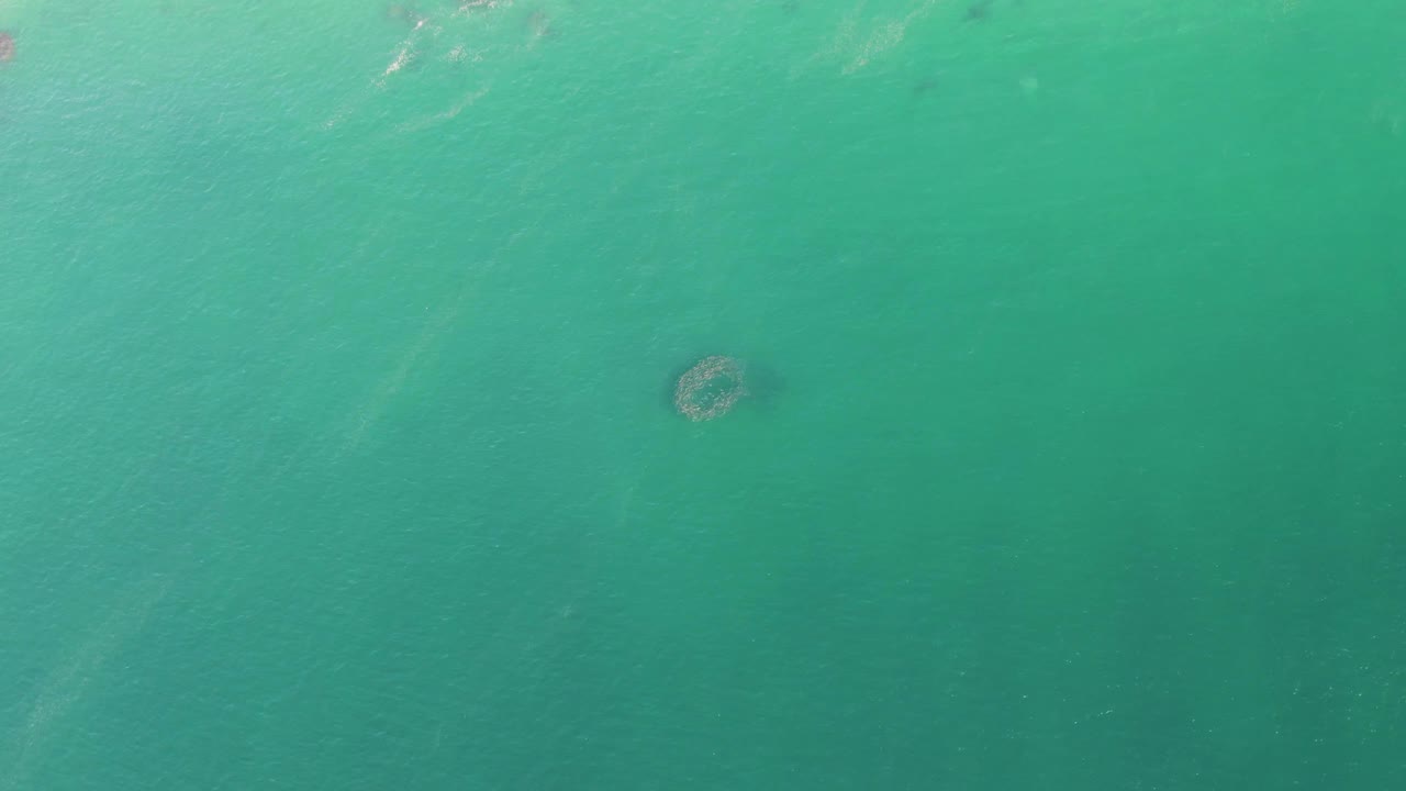 Buzzards Beach aerial frame with the circular underwater structure still centered in green-blue water, with faint ripple streaks crossing the upper part and no visible people.