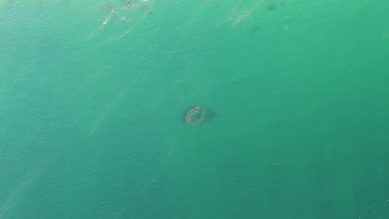 Mid-clip frame from Buzzards Beach showing the circular underwater structure in clear turquoise sea, with darker green shading across the lower water and no visible people.