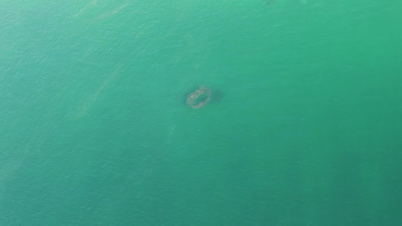 Turquoise ocean water at Buzzards Beach with soft ripples and the small submerged structure held near the upper middle of the overhead frame, with no visible people.