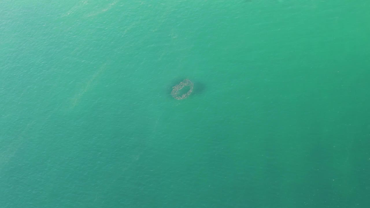 Clear turquoise water in Baja California Sur shows a small reef-like submerged shape near the upper center-right, with faint surface texture and no visible people.