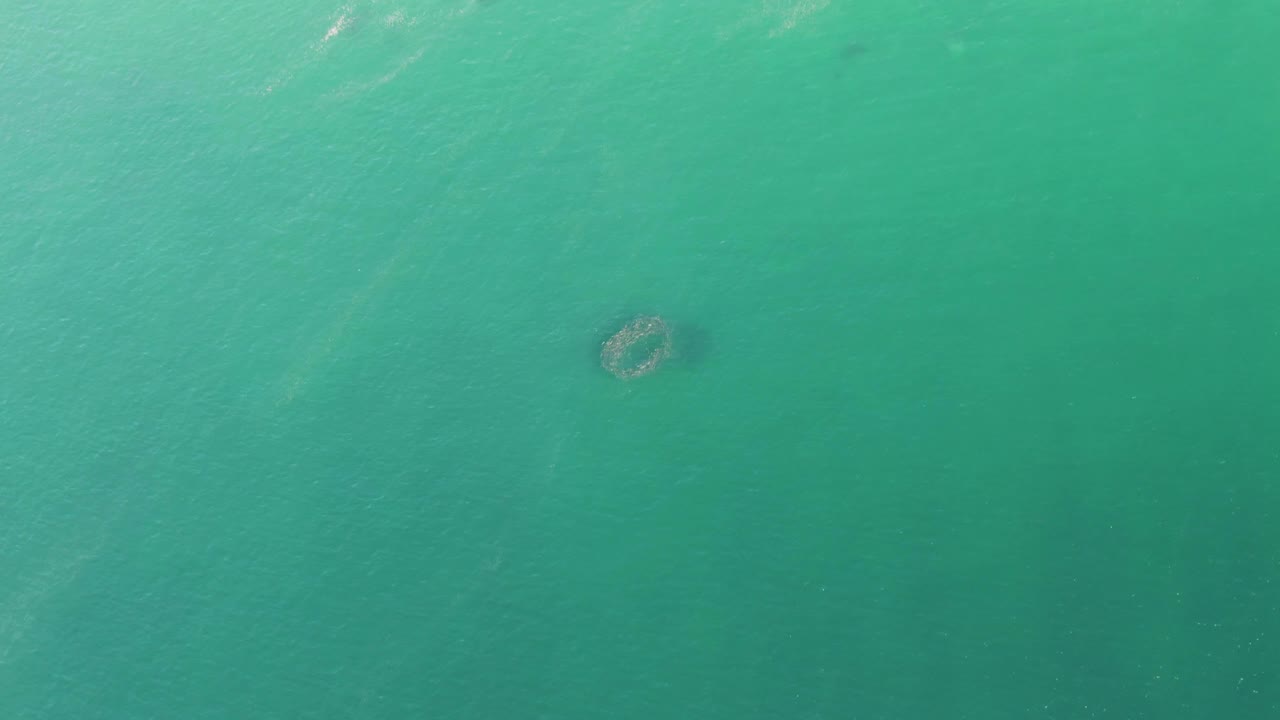 Aerial view over blue-green Buzzards Beach water, showing the circular submerged form right of center with pale surface ripples and no visible people.