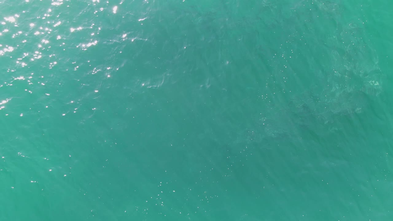 Clear turquoise ocean surface at Buzzards Beach, Baja California Sur, with a cluster of bright glints near the top edge, faint underwater texture, and no visible people.