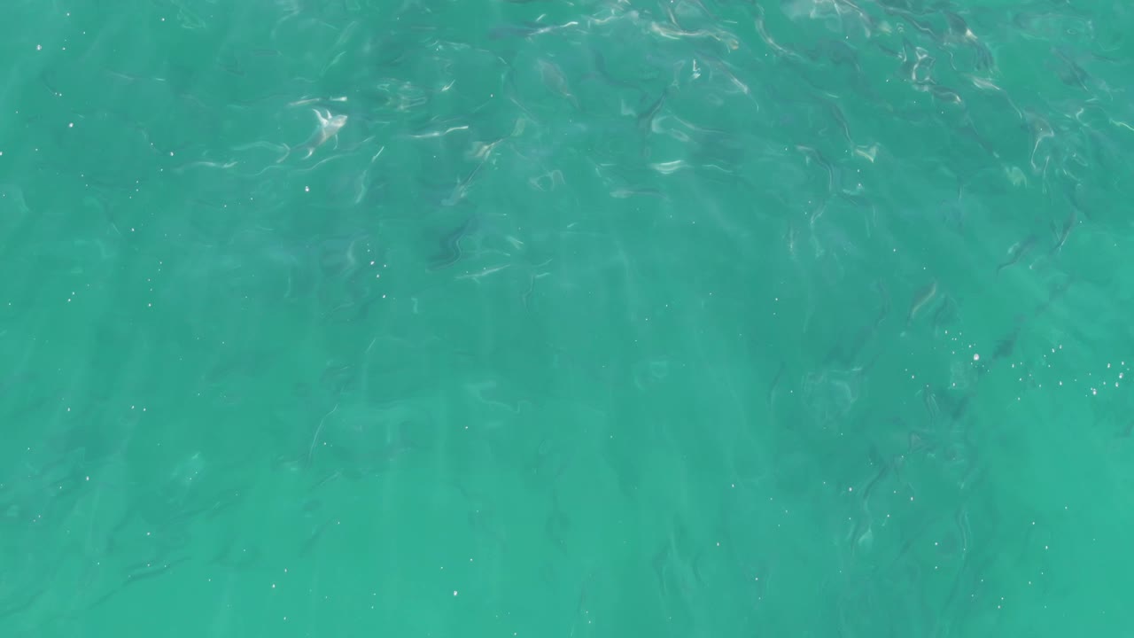 Top-down turquoise sea surface at Buzzards Beach in Baja California Sur includes scattered bright glints, faint underwater texture, and no visible people.