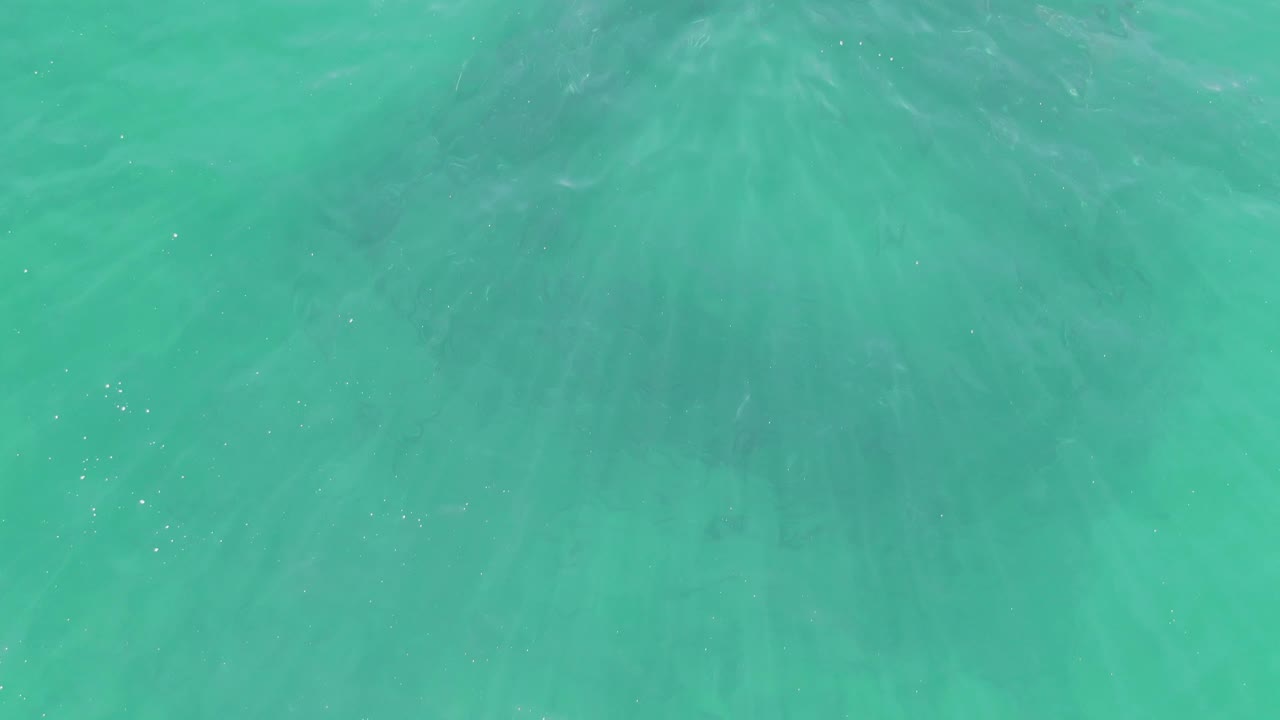 Clear shallow water at Buzzards Beach, Baja California Sur, shows brighter turquoise color, scattered light streaks, subtle darker patches beneath the surface, and no visible people.