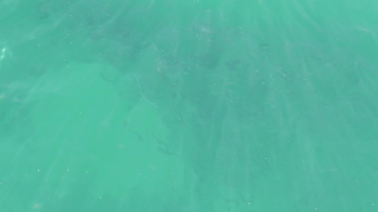 Straight-down view of transparent turquoise sea at Buzzards Beach, with pale sand visible beneath the moving surface and no visible people.