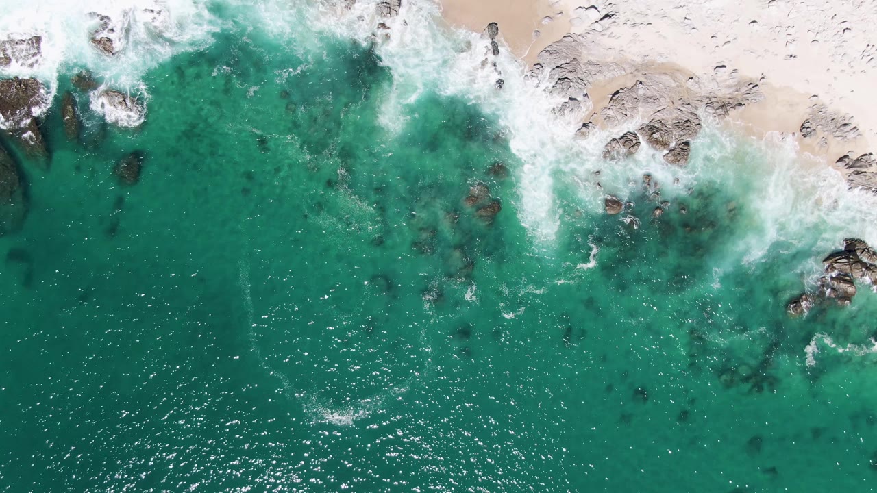 Overhead turquoise surf breaking against rocks and pale sand.