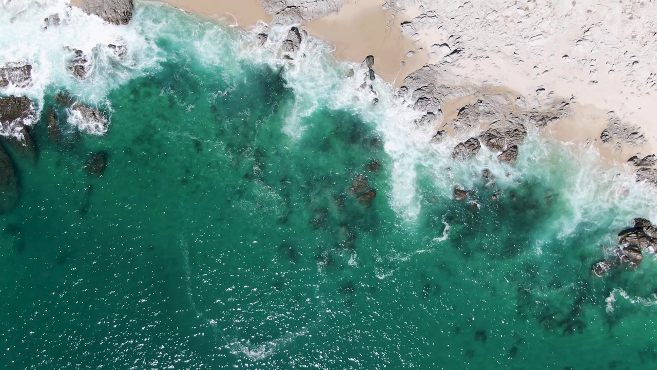 Aerial frame of white waves spreading over dark rocks and pale sand at Buzzards Beach, with turquoise water filling the lower part of the view and no visible people.