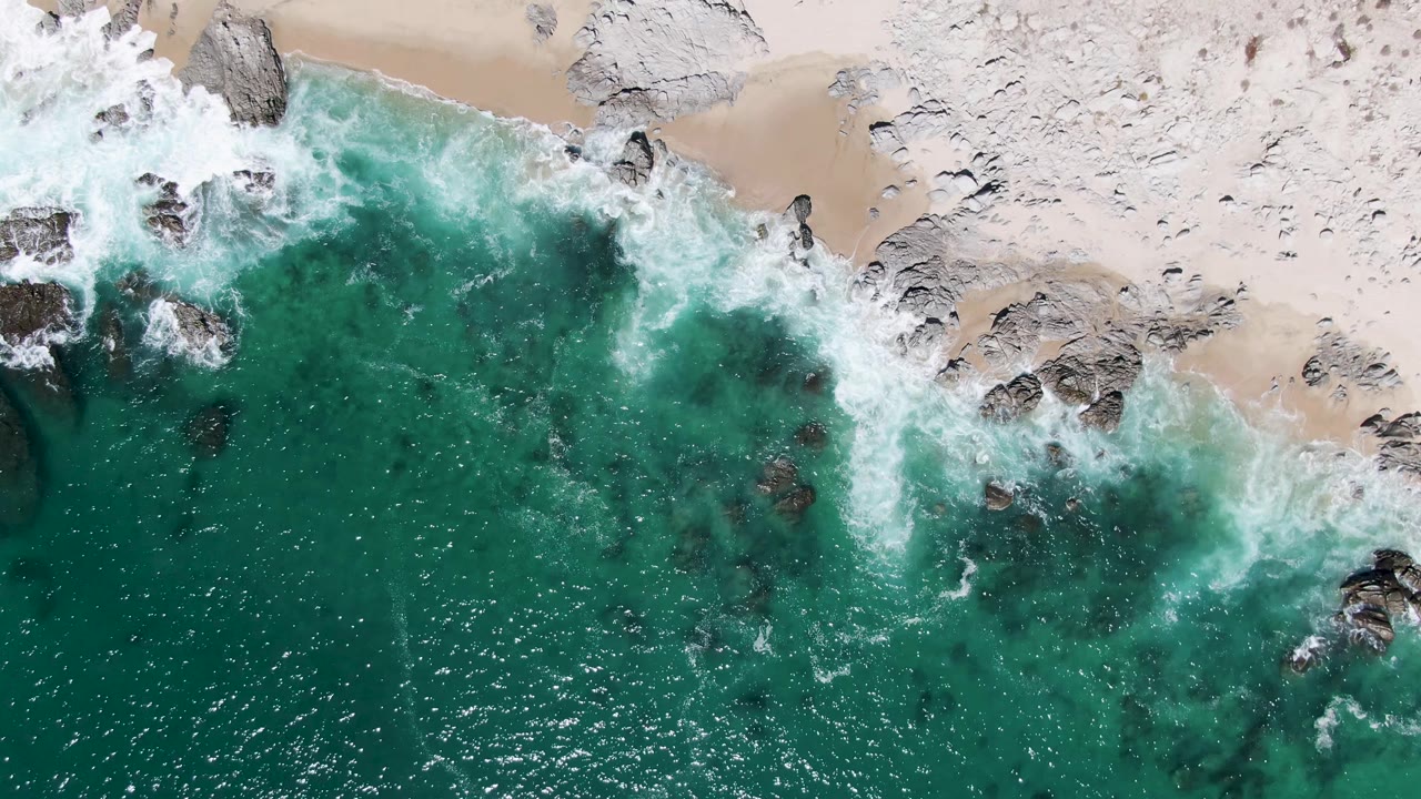 Top-down view of surf curling around scattered shoreline rocks on Buzzards Beach, Baja California Sur, with the sandy beach above and no visible people.