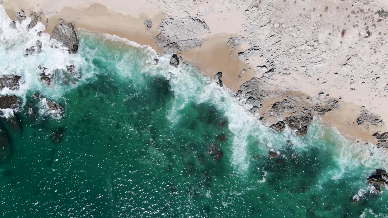 Aerial coastal frame at Buzzards Beach where turquoise ocean meets pale sand and dark rocks under fresh white foam, with no visible people or structures.