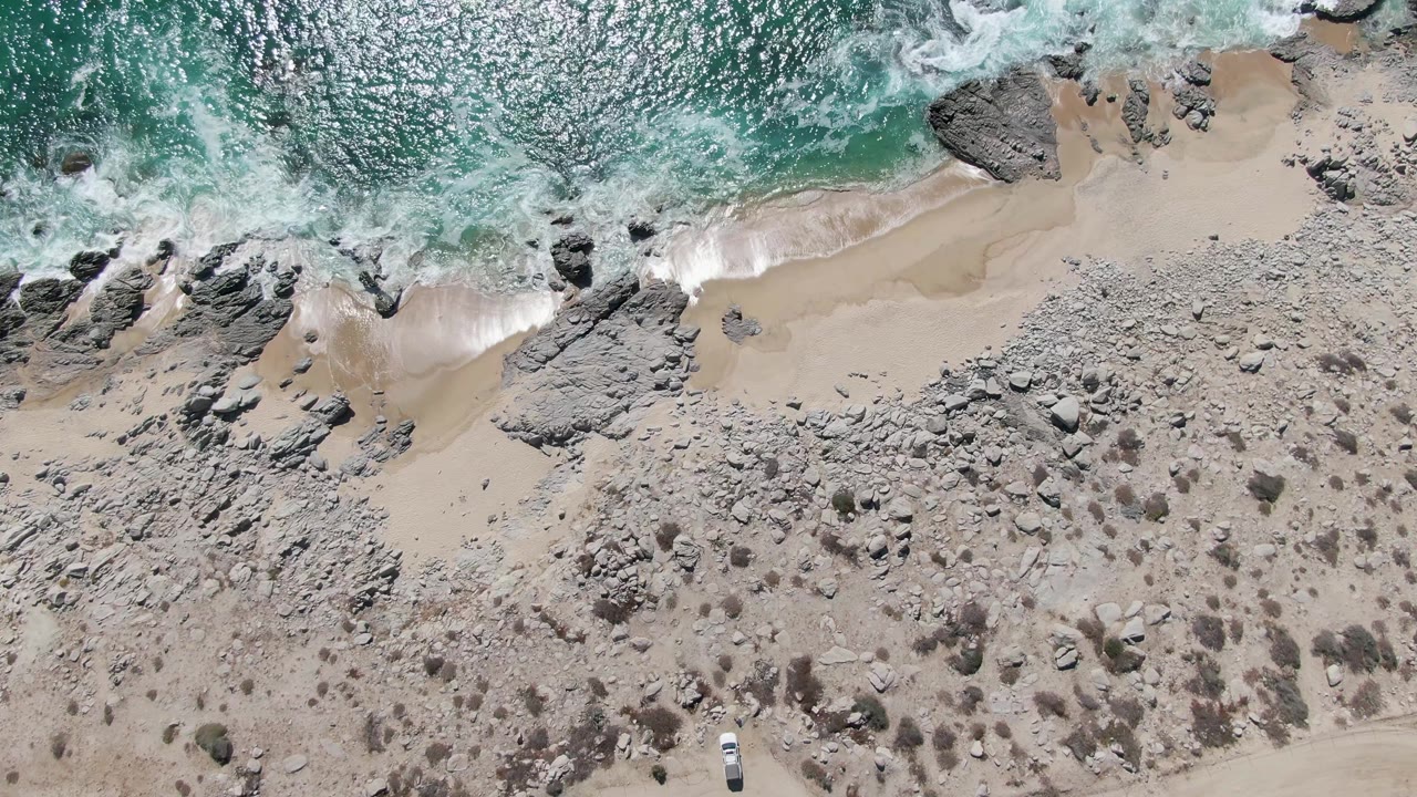 Buzzards Beach aerial frame with a white wave line sliding along the sand, darker rocks clustered near the water, scrub patches inland, and no visible people.