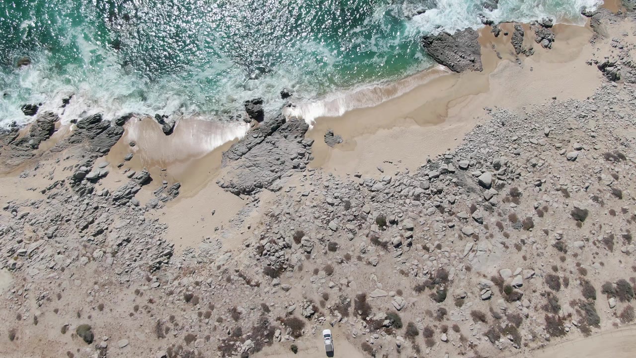 A top-down view at Buzzards Beach shows foamy water pushing into the rocky shoreline, a wider sandy strip, dry scrub-covered ground, and no visible people.