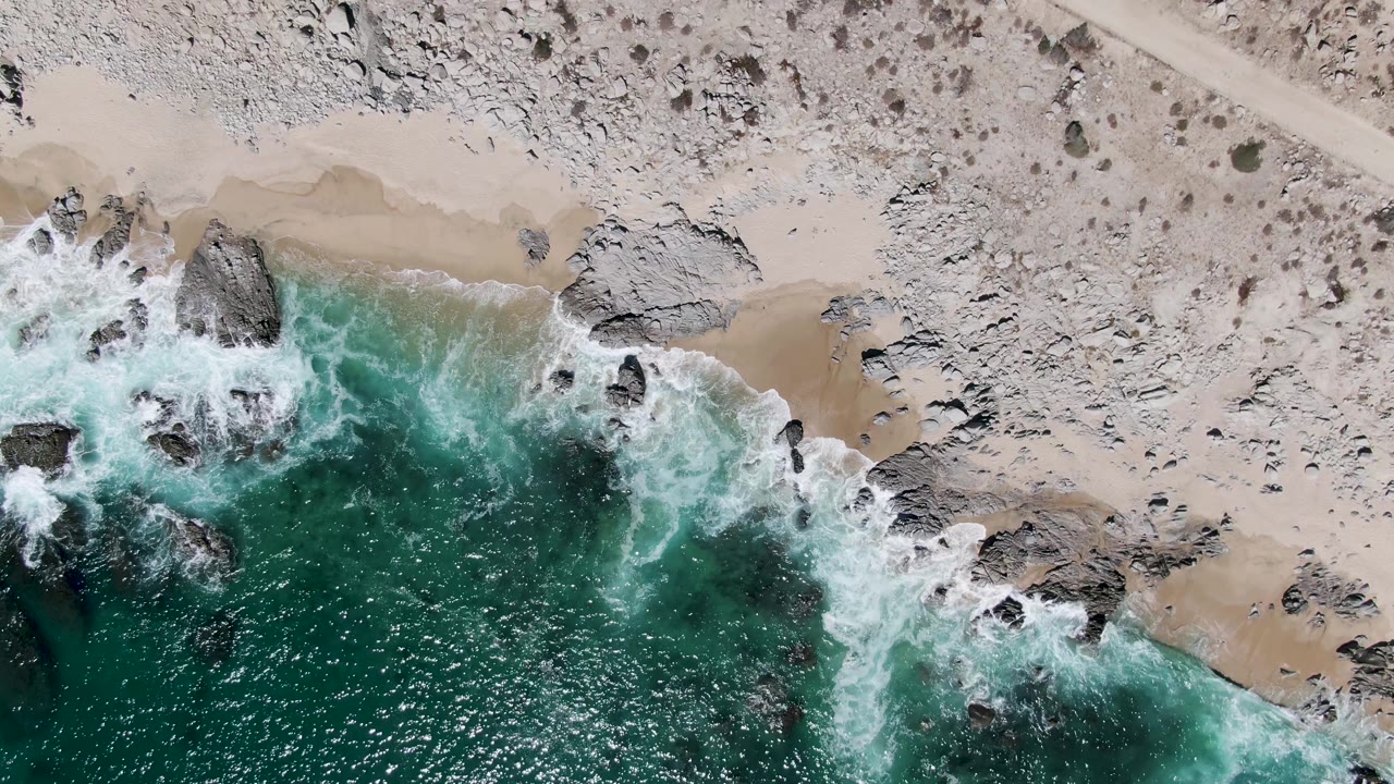 Overhead view of surf, rocks, sand, scrub, dirt road, and a white vehicle.