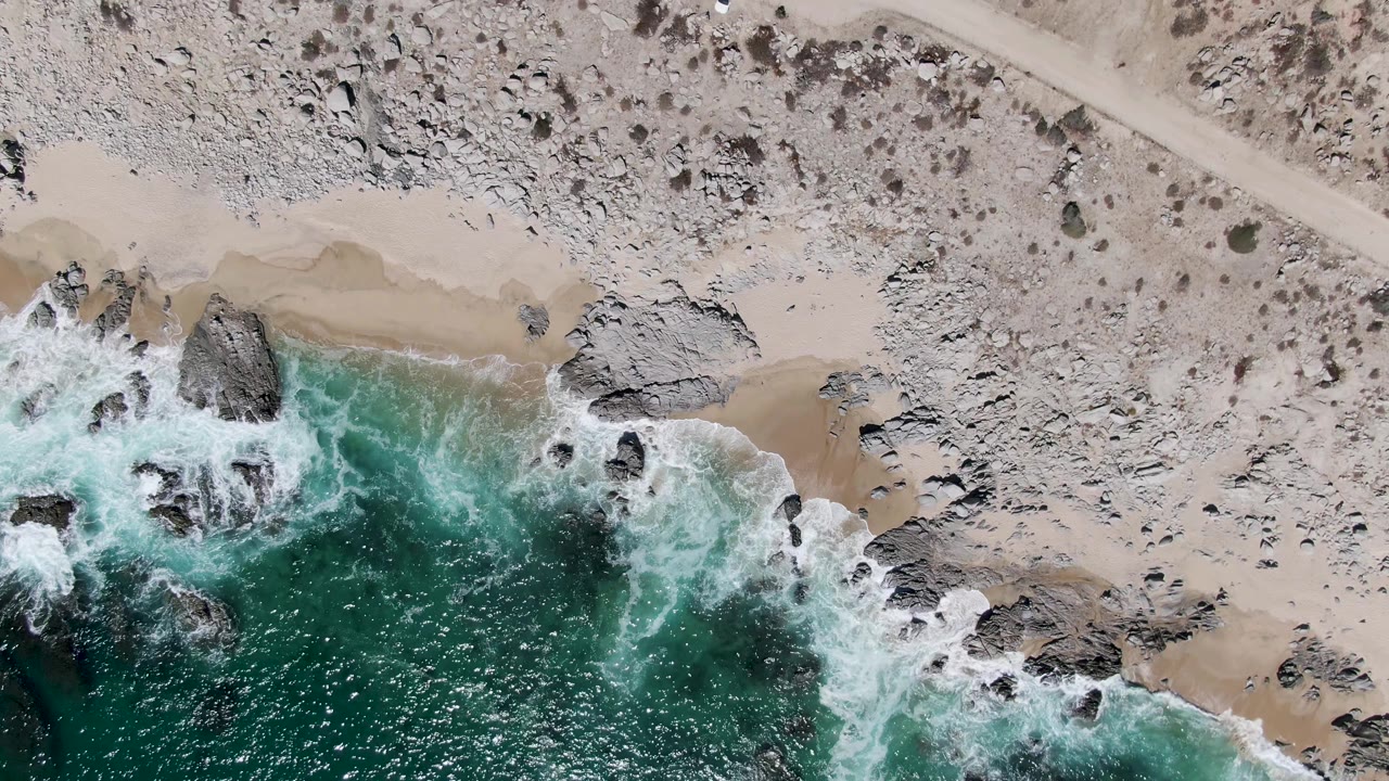 At 0:01, the Buzzards Beach aerial view keeps the white vehicle near the upper road while foamy waves spread around dark rocks and tan sand below, no visible people.