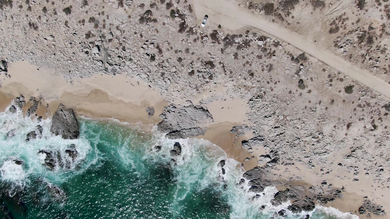 At 0:02, the overhead frame shows the white vehicle farther along the dirt road above a rocky Buzzards Beach shoreline where surf curls into small sandy pockets, no visible people.