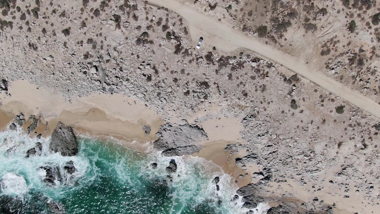 At 0:03, the frame emphasizes the arid scrub and diagonal dirt road above the shoreline, with the white vehicle small in frame and waves breaking over rocks at the lower edge, no visible people.