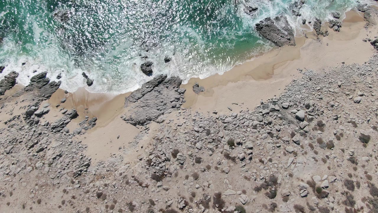Overhead Buzzards Beach frame with bright turquoise water across the top edge, broken white foam over rocks near the surf line, and a broad rocky sandy beach below with no visible people.