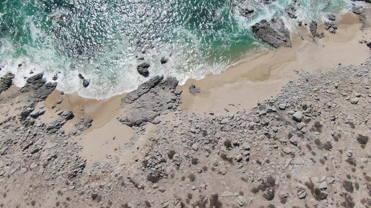 Buzzards Beach appears from above as a fresh wash of white foam curls along the sand, with darker rocks at the waterline and a stony dry beach filling the lower half, with no visible people.