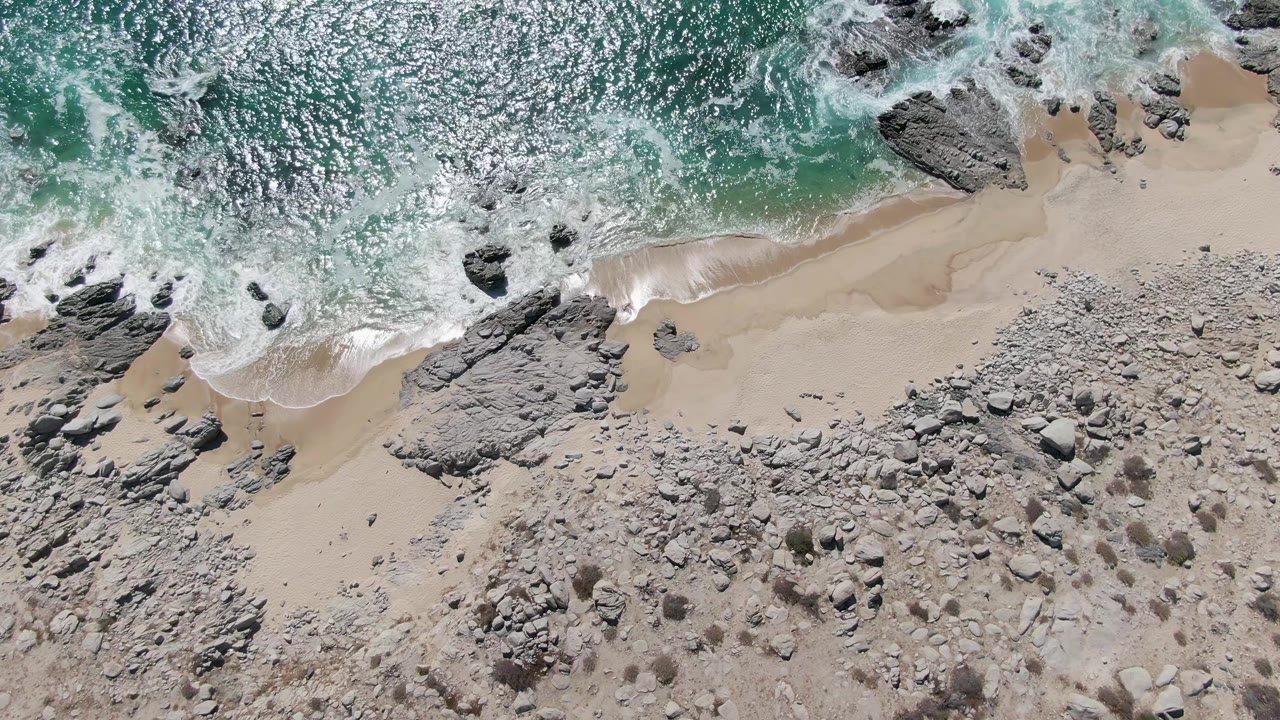Overhead frame of Buzzards Beach surf washing toward pale sand and gray rock shelves, with sparkling turquoise water and no visible people.