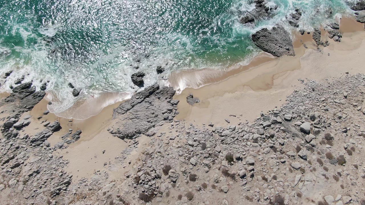 Overhead Buzzards Beach view with a white wave line curling around dark rocks and spreading across the sand, with no visible people.